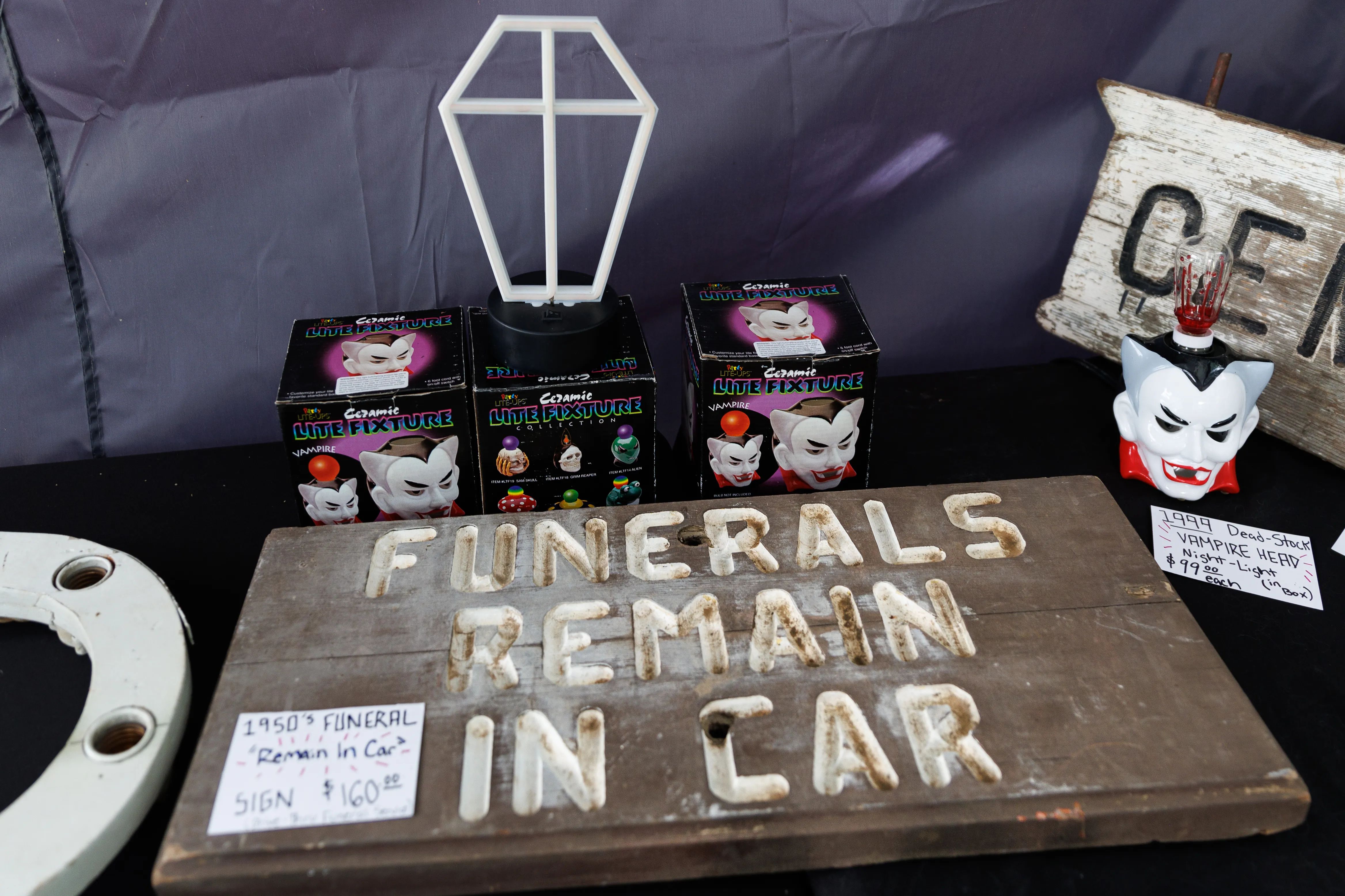 A vintage wood sign that reads "Funerals remain in car" in the foreground, with a coffin shaped light fixture in the background.