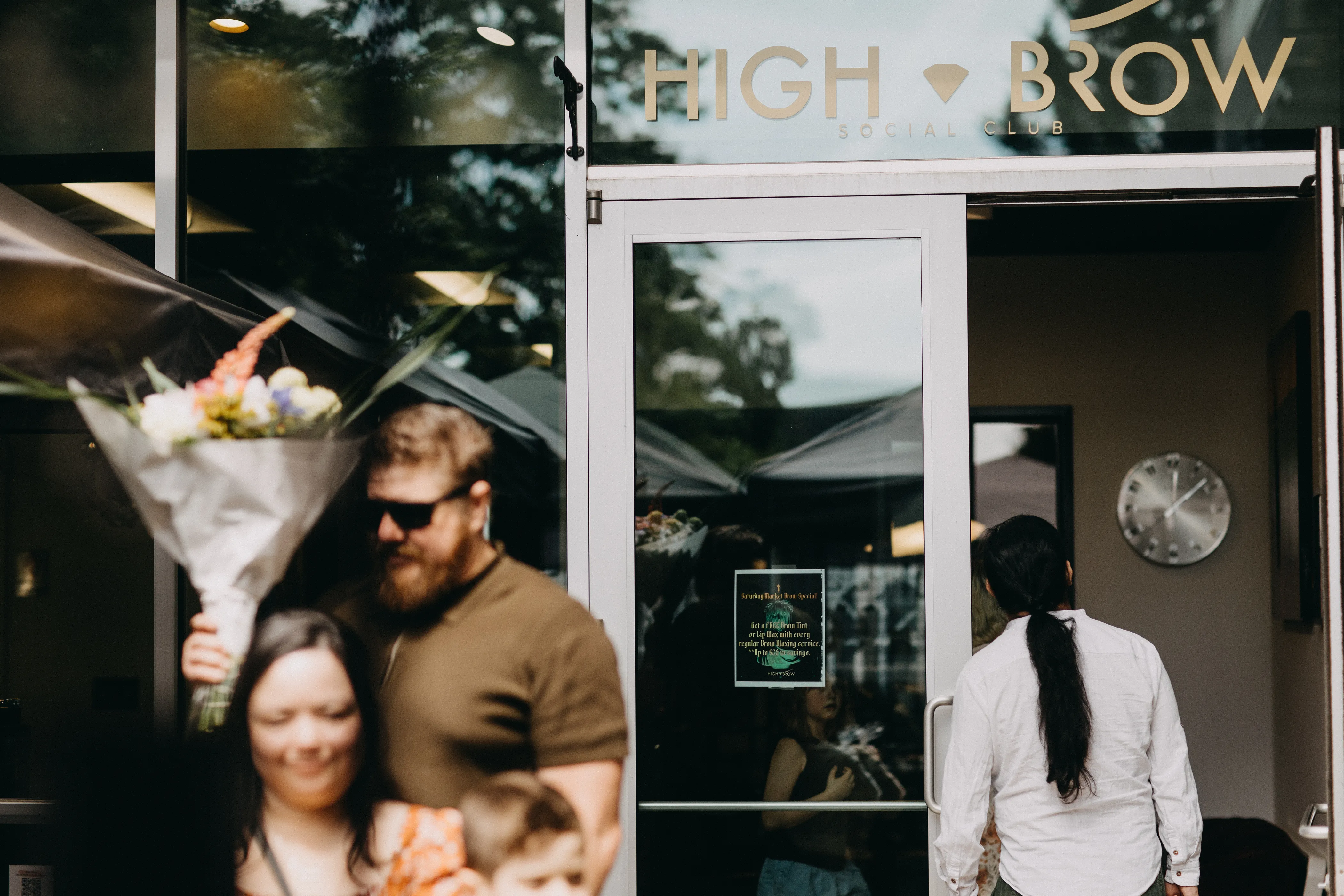 A couple exiting High Brow Social Club after purchasing a bouquet of flowers while a curious passerby peeks in on the Speakeasy Market.