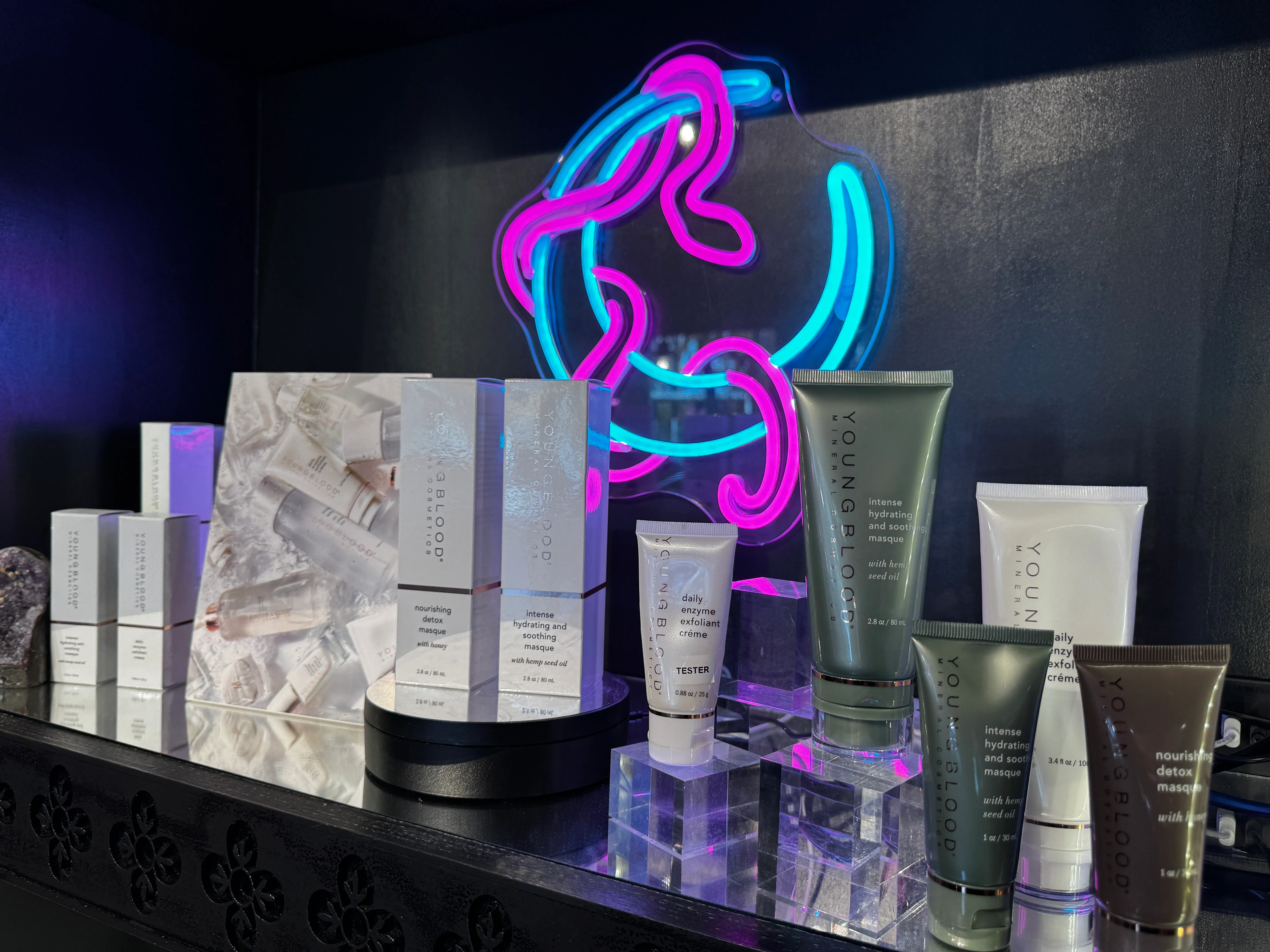 A collection of Youngblood Mineral Cosmetics skincare products, in front of a neon sign featuring a blue crescent moon with a magenta snake loosely coiled around it