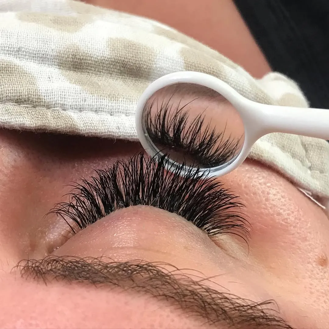 A small mirror being held under a client's lashes for final review of her eyelash work