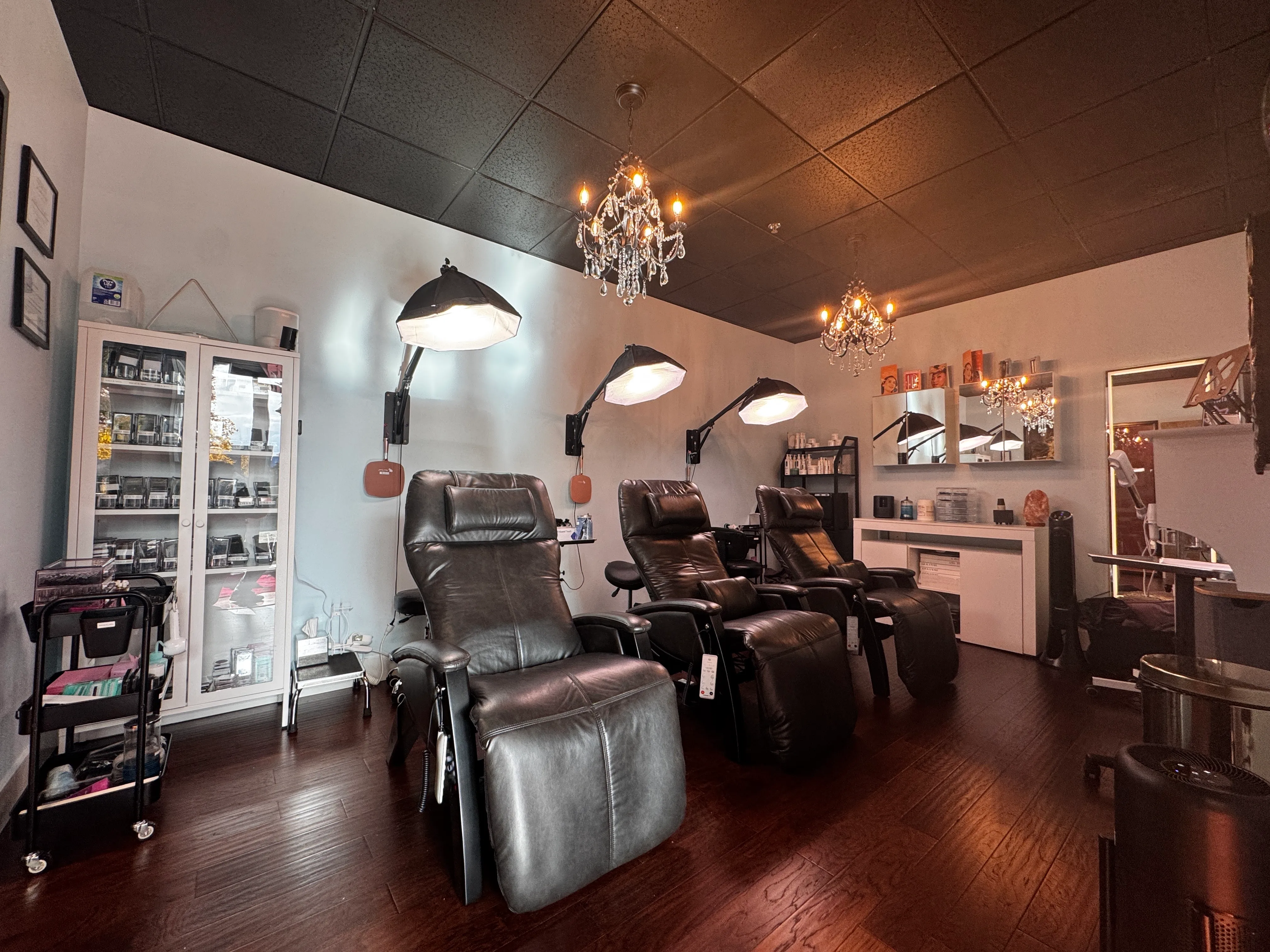 Three plush, black leather electric treatment recliners, above each of which is are articulated professional lights with softbox diffusers, neatly arranged in a modern and upscale beauty tratment studio.